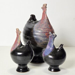 Könnte beinhalten: Drei Keramikvasen in Hühnerform mit schwarzer, blauer und rosa Glasur. Die größte Vase hat einen hohen, spitzen Schnabel und einen runden Körper. Die beiden kleineren Vasen haben kürzere Schnäbel und runde Körper.