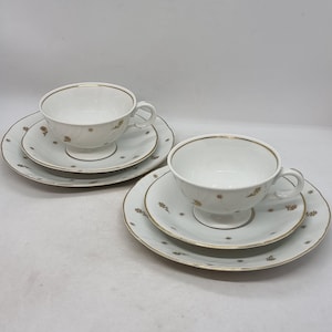 May include: Two white teacups and saucers with a gold trim and a delicate floral pattern. The cups have a footed base and the saucers have a scalloped edge.