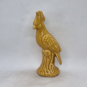 Può includere: Una statuetta in ceramica di un uccello giallo appollaiato su un ramo d'albero. L'uccello ha una testa crestata e piume dettagliate.