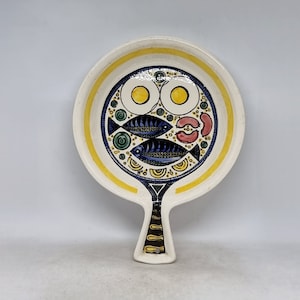 Może przedstawiać: Biała ceramiczna tacka do serwowania z żółtą obwódką i kolorowym wzorem ryb, jajek i innych geometrycznych kształtów. Taca ma uchwyt w kształcie ryby.
