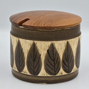 Puede incluir: Un tarro cilíndrico de cerámica con tapa de madera. El tarro es marrón con una banda beige con diseños de hojas marrones oscuras. La tapa de madera tiene una pequeña muesca para una cuchara. Un recipiente decorativo para guardar en la cocina.