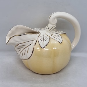 May include: A yellow ceramic pitcher shaped like a pear with gold leaf accents and a white handle.