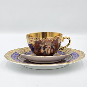 May include: A porcelain teacup and saucer set featuring a deep blue and gold decorative border. The cup displays a detailed historical scene of figures in military attire. The interior of the cup is finished in a reflective gold metallic glaze.