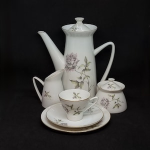 May include: A white porcelain tea set with a floral design. The set includes a teapot, creamer, sugar bowl, and a teacup and saucer. The flowers are pink and green and have a delicate, hand-painted look.