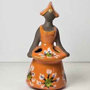 Könnte beinhalten: Orangefarbene Keramikfigur einer Frau mit Hut und Kleid mit weißen und grünen Blumendesigns. Das Kleid hat Löcher zum Aufbewahren von Zahnbürsten oder anderen kleinen Gegenständen.