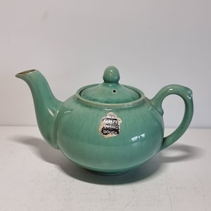 Può includere: Una teiera in ceramica verde con un coperchio e un manico. La teiera ha un'etichetta che recita "KABEERZ OURVAST GOUDA".