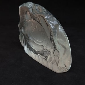 May include: A clear glass sculpture of a sea turtle. The sculpture has a textured surface and a detailed carving of a turtle. The sculpture is on a black surface.