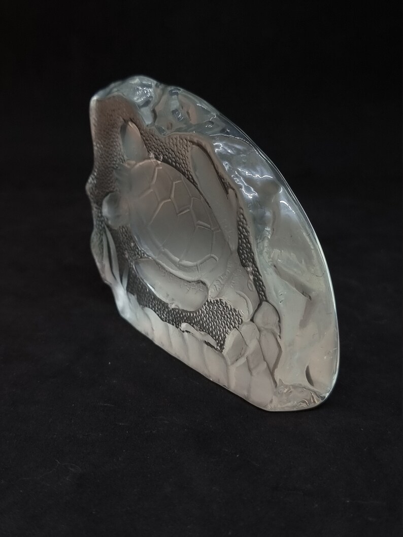 May include: A clear glass sculpture of a sea turtle. The sculpture has a textured surface and a detailed carving of a turtle. The sculpture is on a black surface.