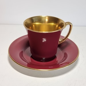 Puede incluir: Un juego de taza y platillo de té de color burdeos y dorado. La taza tiene un borde dorado y el platillo tiene una banda dorada alrededor del borde.