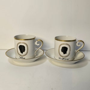 May include: Two white porcelain coffee cups with gold trim and black silhouette portraits. The cup on the left says "Silvia" and the cup on the right says "Carl W. Goetz".