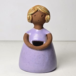 May include: A ceramic figurine of a woman with blonde hair and a purple dress. The figurine has a hole in the front of the dress, making it suitable for holding small items.