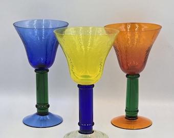 Op de afbeelding: Drie glazen bekers met getextureerde kelken en gladde stelen. De linker beker heeft een blauwe kelk en een groene steel. De middelste beker heeft een gele kelk en een blauwe steel. De rechter beker heeft een oranje kelk en een groene steel.