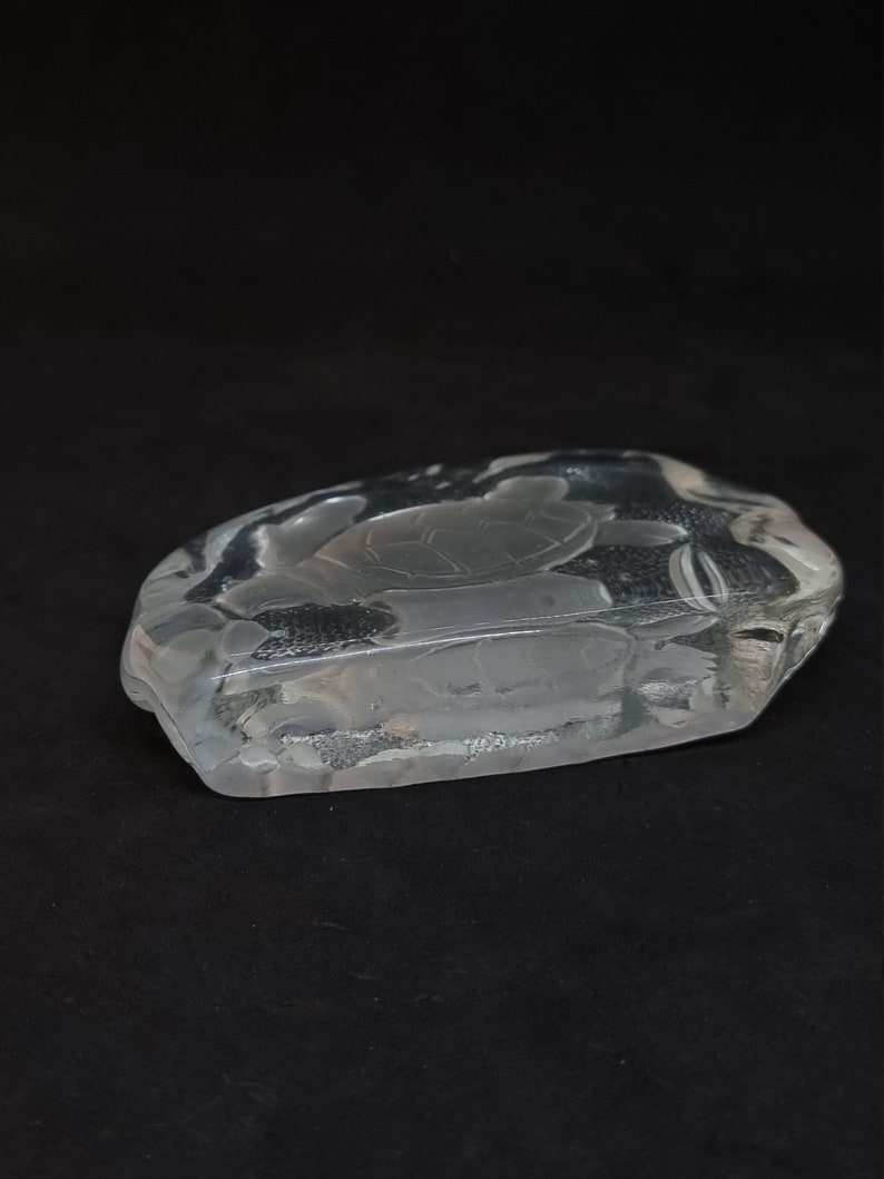 May include: A clear glass ashtray featuring a turtle design. The ashtray is rectangular with rounded edges and a shallow bowl. The turtle is etched into the glass, set against a black background.