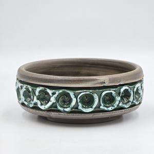 Può includere: Una ciotola in ceramica rotonda con un esterno grigio scuro opaco e un'ampia fascia di smalto verde intenso al centro. La fascia verde è decorata con un motivo ripetitivo di elementi circolari in rilievo a forma di spirale.