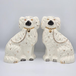 Könnte beinhalten: Ein Paar weißer Keramik-Spaniel-Hundefiguren mit schwarzen Nasen, goldfarbenen Halsbändern mit hängenden Glöckchen und Goldkettendetails. Die Hunde haben texturierte Fellmuster mit kleinen goldenen Akzenten und aufgemalten Augen.