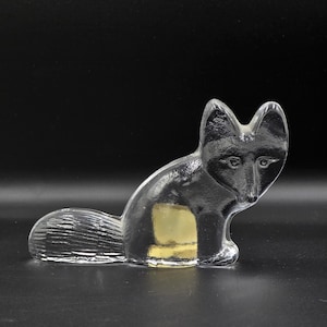 May include: A clear glass fox ornament in a seated position. The fox features a detailed face and tail, with a gold-coloured square on its side. The ornament is set against a black background.