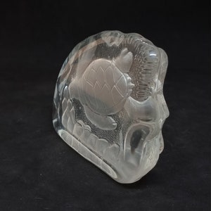 May include: A clear glass sculpture depicting a sea turtle swimming amongst aquatic plants. The sculpture has a rounded shape and is set against a dark background, highlighting the intricate details of the design. The sculpture is made of glass.