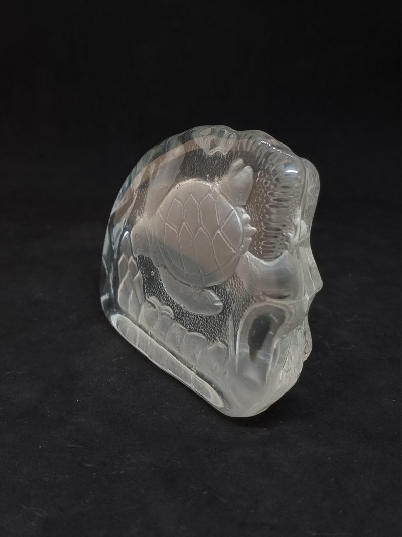 May include: A clear glass sculpture depicting a sea turtle swimming amongst aquatic plants. The sculpture has a rounded shape and is set against a dark background, highlighting the intricate details of the design. The sculpture is made of glass.