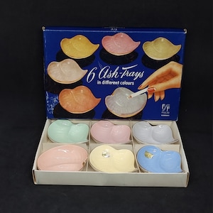 Puede incluir: Un juego de seis ceniceros de cerámica de color pastel en forma de labios. Los ceniceros están dispuestos en una caja con fondo azul y el texto "6 Ash Trays in different colours".