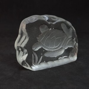 May include: A clear glass sculpture of a sea turtle swimming in the ocean. The turtle is etched into a rectangular block of glass, with details of the shell and flippers visible. The sculpture includes etched seaweed.