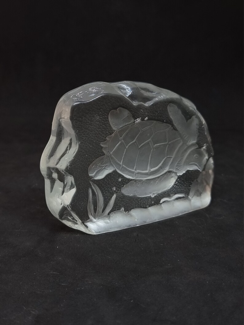May include: A clear glass sculpture of a sea turtle swimming in the ocean. The turtle is etched into a rectangular block of glass, with details of the shell and flippers visible. The sculpture includes etched seaweed.