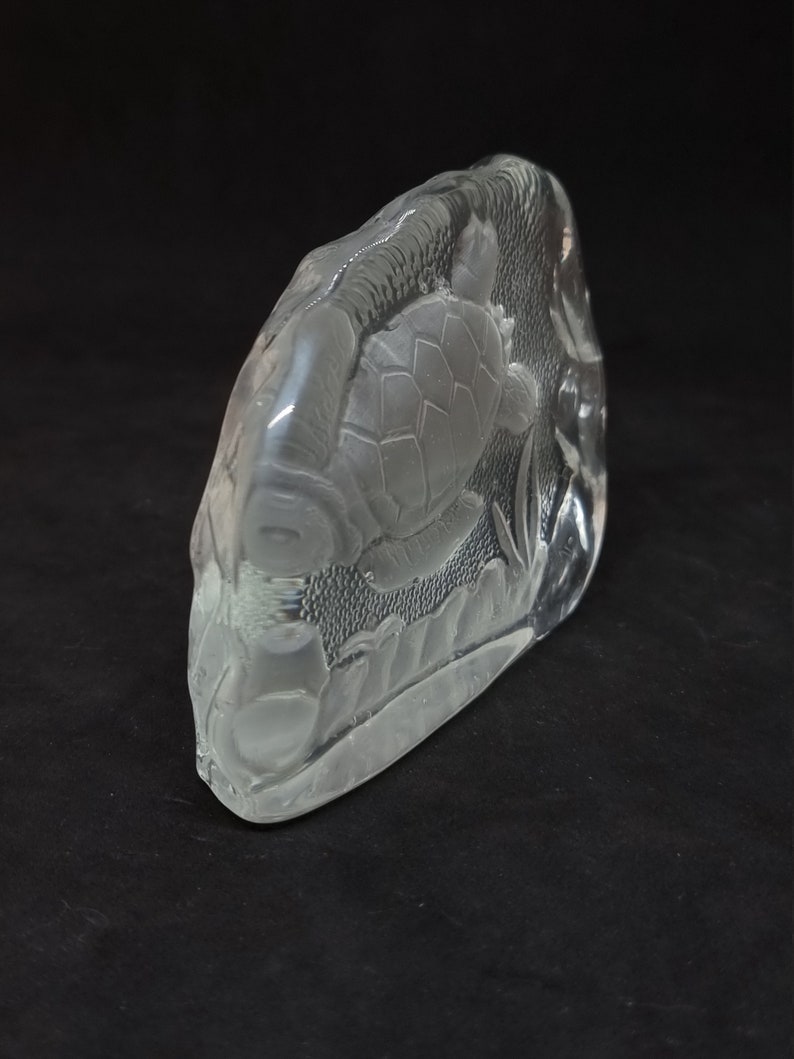 May include: A clear glass sculpture with a detailed relief of a turtle swimming amongst aquatic plants. The sculpture has a rounded, irregular shape against a dark background. The turtle's shell and the plants are intricately carved.