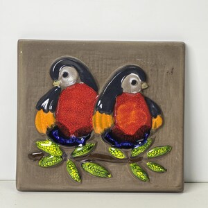 Puede incluir: Dos pájaros de cerámica con detalles rojos, naranjas, azules y negros posados en una rama con hojas verdes. Los pájaros están en relieve sobre un fondo marrón.