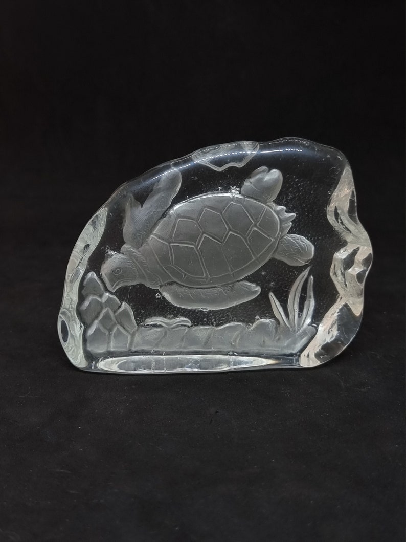 May include: A clear glass sculpture of a swimming sea turtle. The turtle is etched into a rectangular block of glass, with details of the shell, flippers, and aquatic plants. The sculpture is set against a black background.