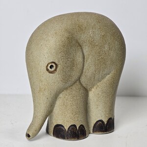 May include: A ceramic elephant figurine in a light tan color with dark brown accents. The elephant has a rounded body, a trunk, and small, dark brown feet. The eye is a simple brown circle.