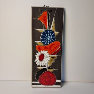May include: A rectangular ceramic tile with a black background and a colorful abstract design. The design features a white flower, red and orange shapes, and a blue and white patterned circle.