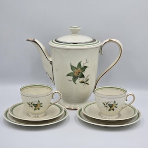 Puede incluir: Un juego de té de porcelana color crema que incluye una tetera alta con tapa, dos tazas y dos platillos. Cada pieza está decorada con un motivo floral verde y amarillo, bandas verdes finas y bordes dorados en los bordes y las asas.