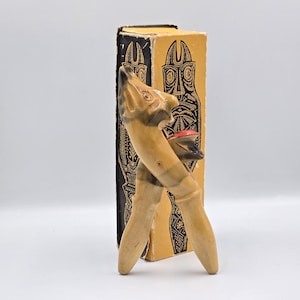 Op de afbeelding: Een houten notenkraker in de vorm van een gestileerde figuur met een rode mond. De notenkraker is lichtbruin met donkerbruine accenten. Hij staat voor een doos met zwarte en gouden tribale ontwerpen.