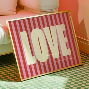 May include: A framed artwork with the word "LOVE" in large, white capital letters. The background features alternating vertical stripes of red and pink. The frame is gold-colored, and the artwork is leaning against a white couch with pink pillows.