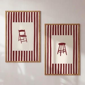May include: Two framed art prints featuring illustrations of furniture. One print shows a red chair, and the other shows a red stool. Both prints have a cream-colored center with a red and white striped border and a wooden frame.