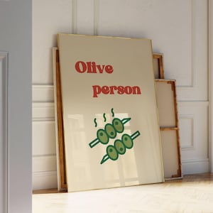 Op de afbeelding: Een ingelijste kunstdruk met de woorden "Olive person" in het rood, boven een afbeelding van twee olijfspiesen. De achtergrond is lichtbeige en de lijst heeft een gouden rand. De kunstdruk leunt tegen een witte muur.