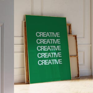 May include: A large green poster with the word "CREATIVE" repeated five times in white. The poster has a gold frame and leans against a white wall. Other framed canvases are behind it.
