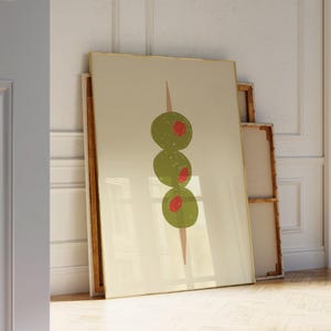 May include: A framed art print depicting three green olives with red centres on a cocktail stick. The artwork has a minimalist style with a beige background and a gold frame. The print is leaning against a white wall.
