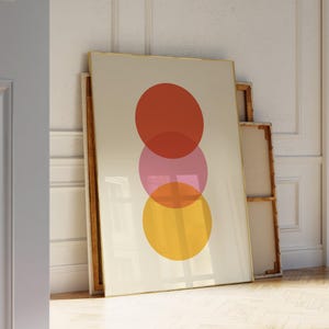 May include: A framed art print featuring three overlapping circles in shades of red, pink, and yellow against a cream background. The artwork has a gold frame and is leaning against a wall.