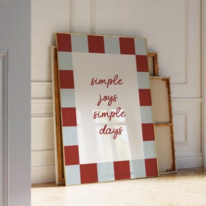 Może przedstawiać: Duże oprawione dzieło sztuki ze złotą obwódką. Grafika przedstawia białe tło ze słowami "simple joys simple days" w kursywie. Rama ma czerwono-niebieski wzór w kratkę.
