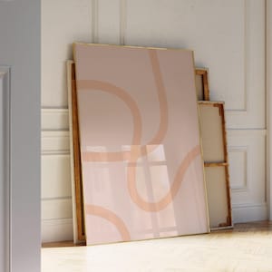 May include: A large framed artwork with a gold border leans against a wall. The artwork features a blush pink background with abstract peach-colored curved lines. Additional unframed canvases are stacked behind it, creating a layered display.