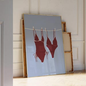 Pode incluir: Impressão emoldurada com dois fatos de banho vermelhos pendurados num varal contra um fundo azul claro. Os fatos de banho são representados com desenhos de linhas simples, e a moldura tem uma borda dourada. A obra de arte está encostada a uma parede.