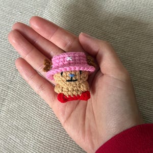 Peut inclure: Petite figurine au crochet représentant un personnage de dessin animé, tenue dans une main. Le personnage a un corps beige, un chapeau rose avec une étoile blanche, des bois bruns et un nœud papillon rouge. La figurine mesure environ 5 cm de haut.