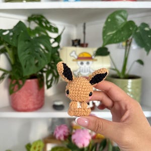 Può includere: Una piccola figura amigurumi all'uncinetto di colore marrone chiaro che ricorda una creatura simile a una volpe, con grandi orecchie a punta con dettagli interni neri, piccoli occhi neri e una macchia bianca sul petto.