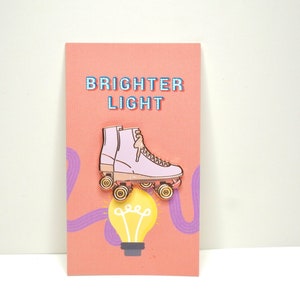May include: Pink roller skate enamel pin with gold wheels and a light bulb graphic. The pin says "Brighter Light" on the card.