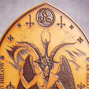May include: A gold-colored metal plaque with a raised image of a horned figure with wings. The figure has a pentagram on its forehead and is surrounded by stars and crosses. The text "As Above, So Below" is inscribed on the plaque.