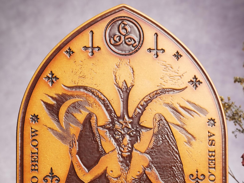 May include: A gold-colored metal plaque with a raised image of a horned figure with wings. The figure has a pentagram on its forehead and is surrounded by stars and crosses. The text "As Above, So Below" is inscribed on the plaque.