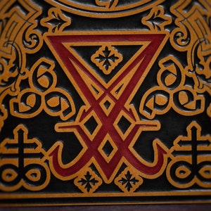 May include: A close-up of a black leather-bound book with a red and gold embossed design. The design features a stylised symbol with a cross and a circle, surrounded by intricate floral patterns.