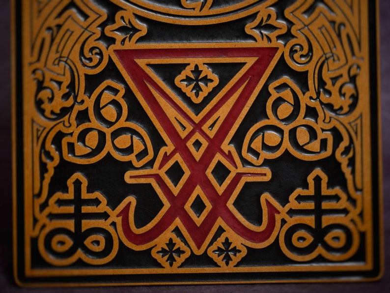 May include: A close-up of a black leather-bound book with a red and gold embossed design. The design features a stylised symbol with a cross and a circle, surrounded by intricate floral patterns.