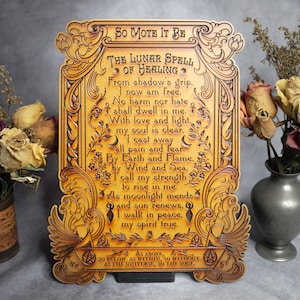 Może przedstawiać: Drewniana tablica z ozdobnymi rzeźbami i tekstem "SO MOTE IT BE" i "THE LUNAR SPELL OF HEALING". Tablica ma ozdobną ramę i jest eksponowana z suszonymi kwiatami w wazonach. Tablica ma około 25 cm wysokości.