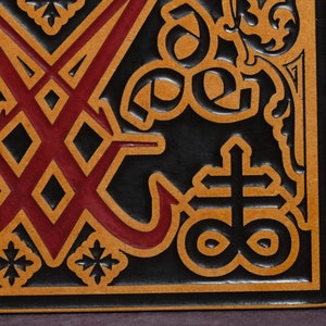 May include: A black leather square with a red and brown design. The design features a cross with a circle around it, and a symbol with two intersecting lines.
